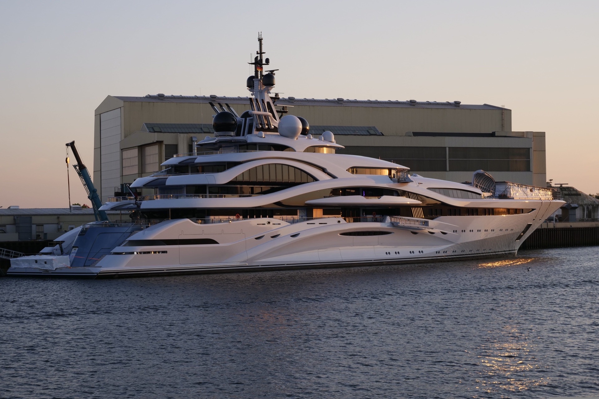 Un superyacht ormeggiato davanti al capannone di un cantiere
                      navale, nella luce bassa del tramonto.